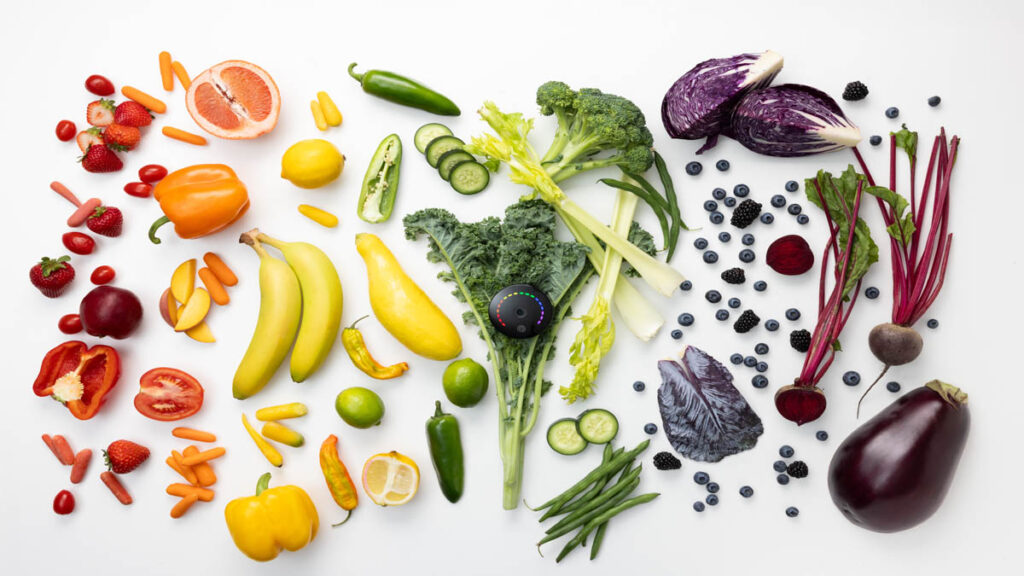 Frutas y verduras frescas junto al dispositivo Prysm iO sobre una mesa.